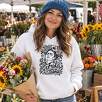 A woman at an outdoor flower market wearing a white hoodie with a black graphic of Frida Kahlo and the text 'Oh Frida GIVE ME COURAGE', paired with a blue knit beanie and holding a bouquet of sunflowers and wildflowers.