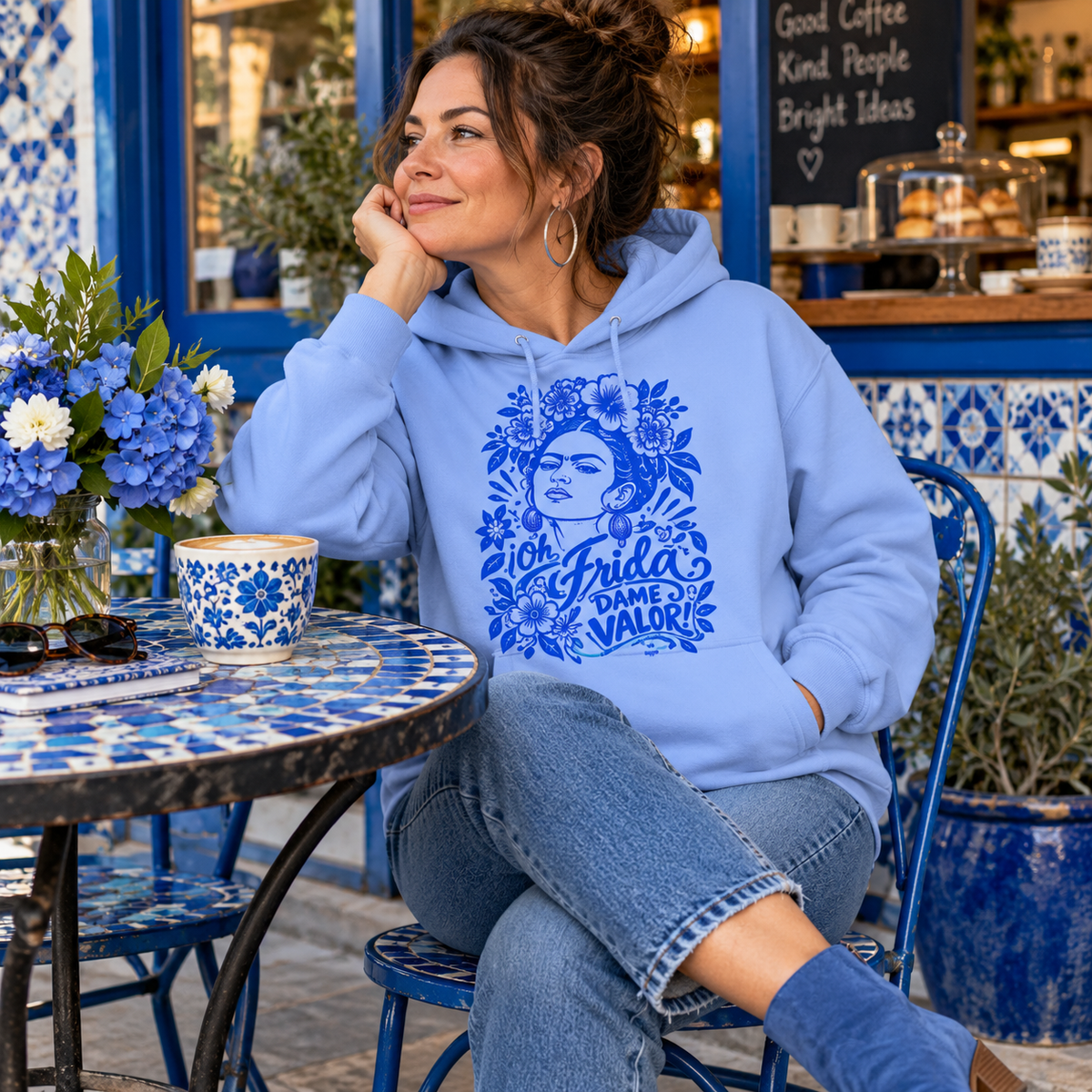 A woman sits at a blue-and-white tiled cafe table wearing a light blue hoodie with a blue graphic print of Frida Kahlo and the text in spanish: Oh Frida Dame Valor.