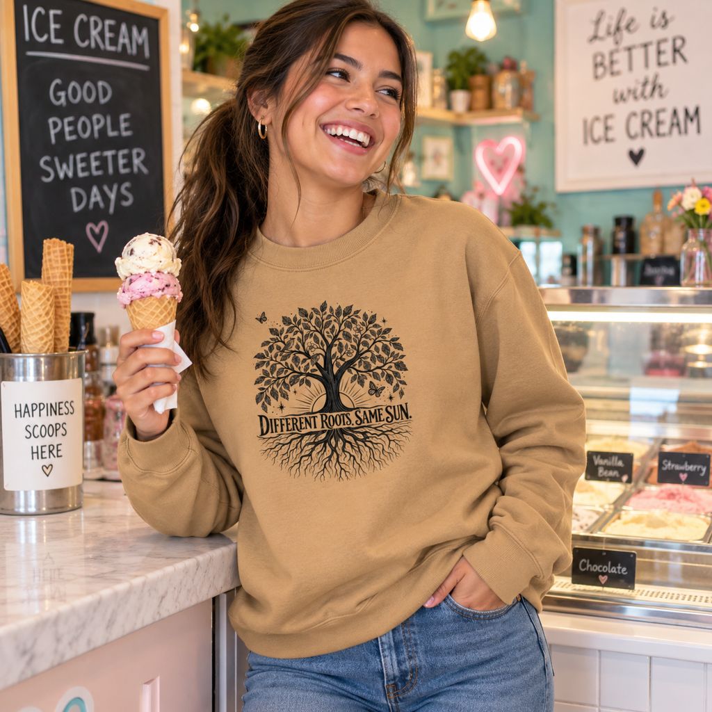 A woman in a cafe setting wearing a tan crewneck sweatshirt with a black graphic of a tree and the text 'Different Roots Same Sun'. She is holding an ice cream cone.