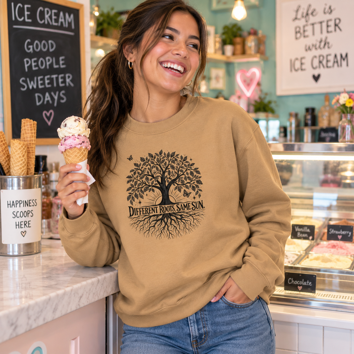 A woman in a cafe setting wearing a tan crewneck sweatshirt with a black graphic of a tree and the text 'Different Roots Same Sun'. She is holding an ice cream cone.