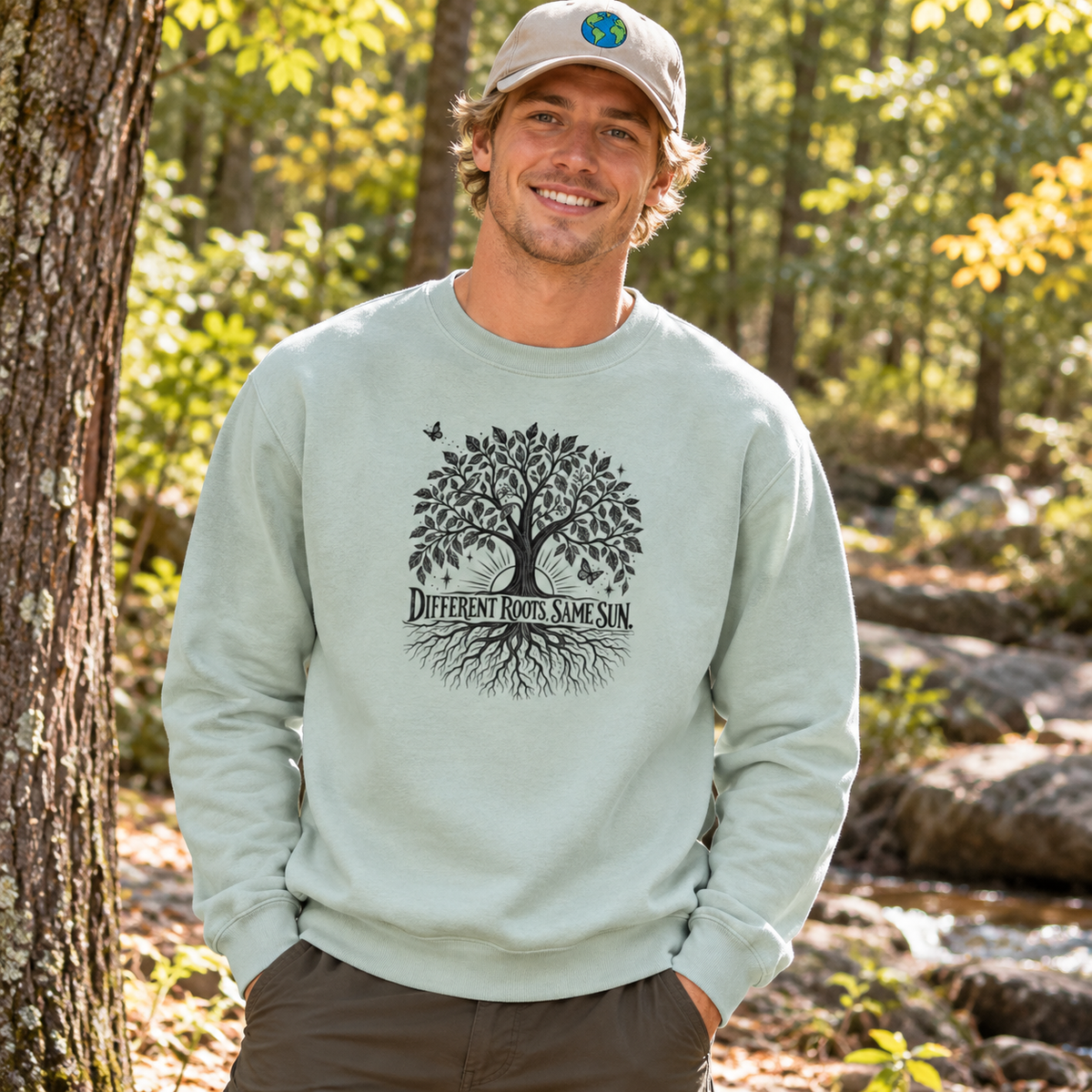 A man standing outdoors in a wooded area wearing a light sage green crewneck sweatshirt with a black graphic of a tree and the text 'Different Roots Same Sun' on the front, paired with a beige baseball cap and brown pants.