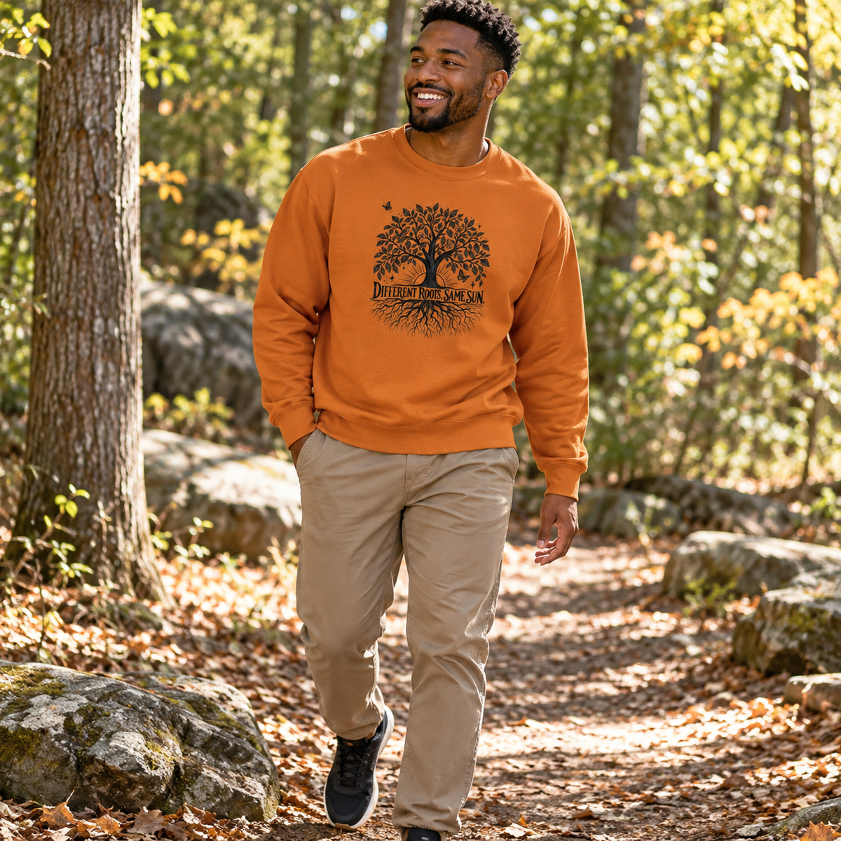 A man walking on an outdoor forest path wearing an orange crewneck sweatshirt with a black tree graphic and the words 'Different Roots, Same Sun" on the front, paired with khaki pants and black sneakers.
