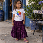 A young girl stands outdoors wearing a white graphic T-shirt with a rainbow wheel and the text 'KINDNESS is my favorite color,' paired with a long purple velvet tiered skirt and pink flip-flops. The background shows a rustic blue doorway, potted plants, and a tiled bench.