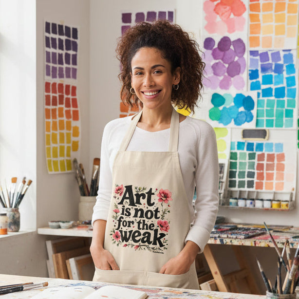 A woman modeling a beige apron with a graphic print that reads 'The Art is not for the weak' in black text with red and green floral accents. She is wearing a white long-sleeve shirt and gray jeans against a plain white background.
