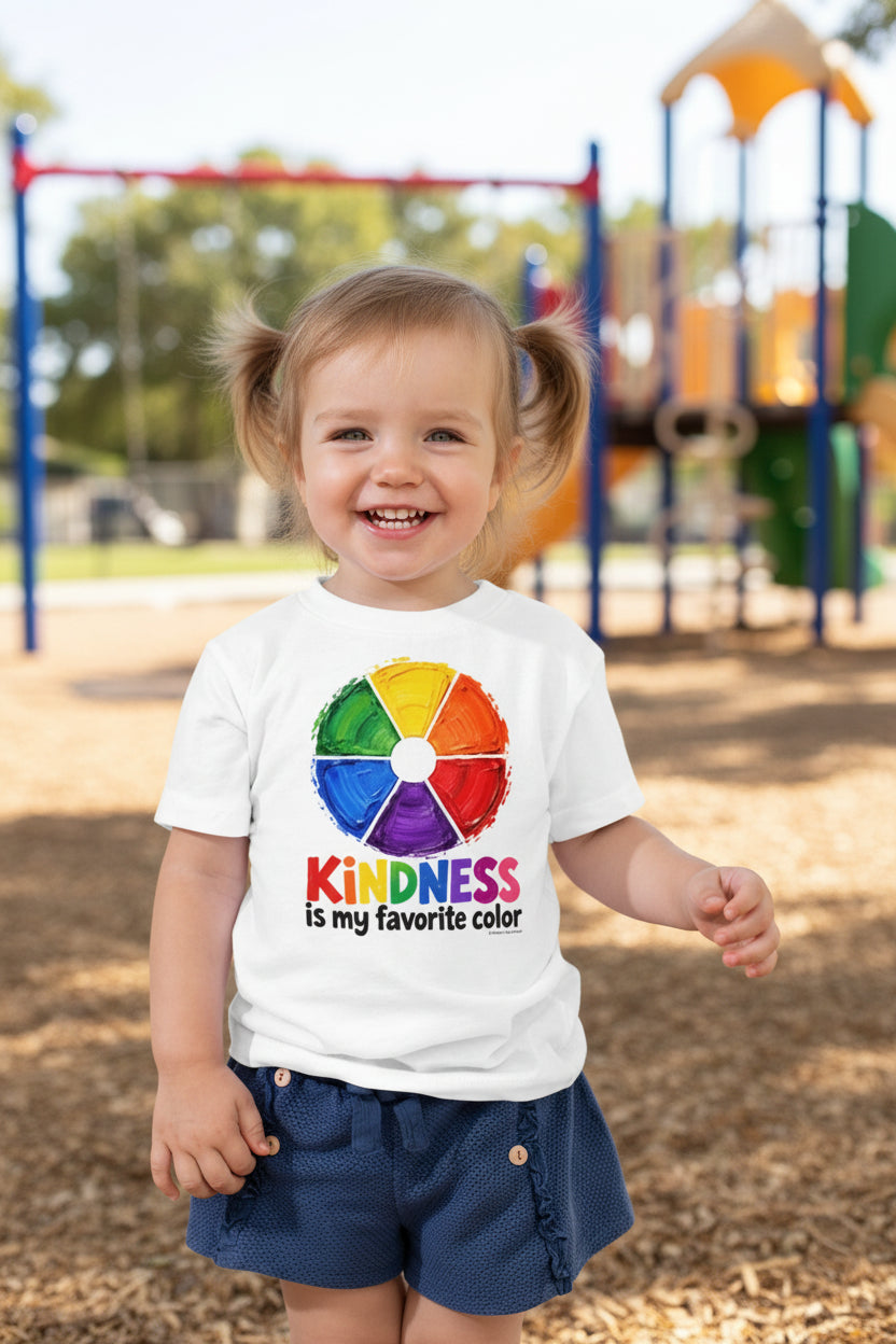 A toddler standing against a playground background wearing a white short-sleeve T-shirt with a colorful rainbow wheel graphic and the text 'Kindness is my favorite color,' paired with dark blue shorts.