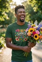A man outdoors holding a large bouquet of colorful flowers, wearing a dark green graphic T-shirt with the text 'KINDNESS IS AN ACT OF IMAGINATION' surrounded by leaves and sunshine.