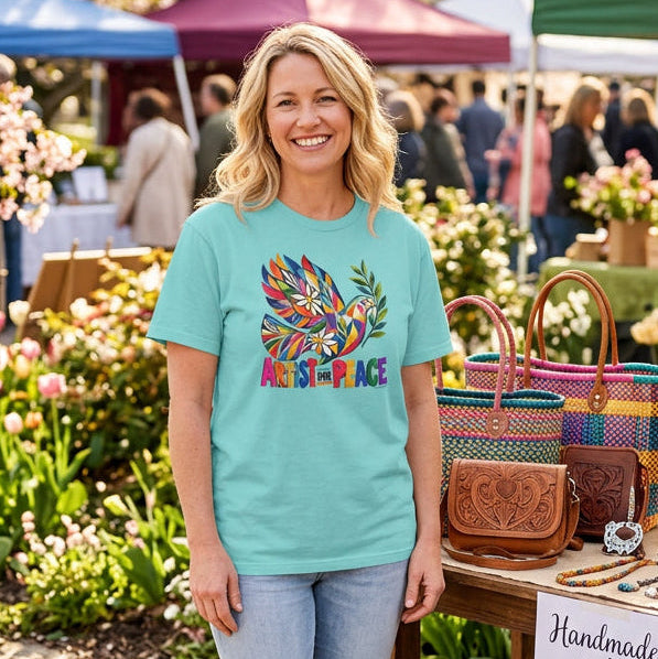 A woman modeling a light blue t-shirt with a colorful graphic of a dove made of geometric shapes and the words 'ARTIST FOR PEACE' on the front, set against a farmers market background and a bag booth.