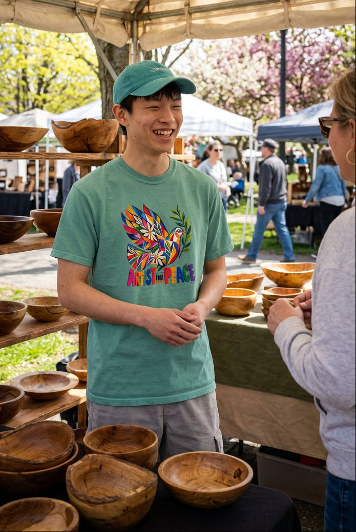 A man wearing a sage green t-shirt with a colorful peace dove graphic and the words 'ARTIST for PEACE' printed on the chest, paired with light gray cargo pants, at an art show