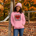 A smiling woman with curly dark hair wears a pink pullover hoodie featuring a colorful heart graphic with the words 'Kindness is Free' in black script. She is posing with her hands in the front pockets of the hoodie against a solid white background.