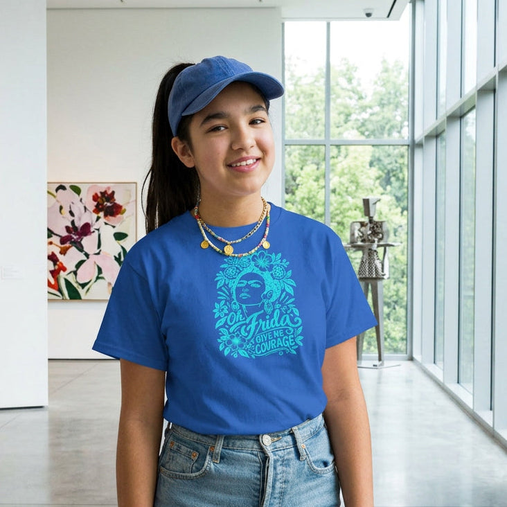 A young girl with long dark hair stands against a solid tan studio background, wearing a bright blue graphic t-shirt with a turquoise illustration of a woman's face and the text 'Oh Frida Give me Courage'. She is also wearing blue jeans and layered gold necklaces.