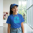 A young girl with long dark hair stands against a solid tan studio background, wearing a bright blue graphic t-shirt with a turquoise illustration of a woman's face and the text 'Oh Frida Give me Courage'. She is also wearing blue jeans and layered gold necklaces.