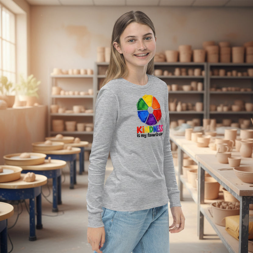 A young woman modeling a light heather gray long-sleeve t-shirt with a colorful circular graphic and the text 'KINDNESS is my favorite color!' printed on the chest, paired with light blue jeans against a pottery studio background